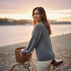 Woman in a gray sweater standing next to a bicycle with a basket by a waterfront at sunset.