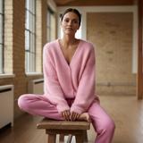 Woman wearing a pink outfit sitting on a wooden stool in a room with large windows.