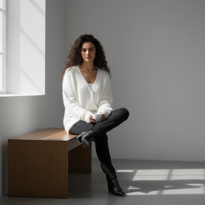 Woman sitting on a wooden bench holding a cup in a minimalistic room with a window.