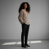 Woman wearing a beige sweater and black pants standing in a softly lit room.