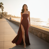Woman in a burgundy strapless gown with a high slit standing on a boardwalk by the ocean.