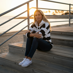 Woman sitting on wooden steps by a waterfront at sunset, wearing a black and white striped sweater and jeans.
