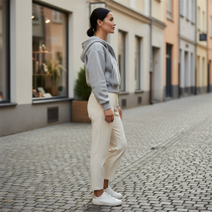 Woman in gray hoodie and white pants standing on a cobblestone street.