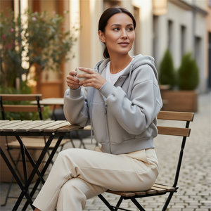 Woman sitting outdoors holding a cup, wearing a gray hoodie and beige pants.