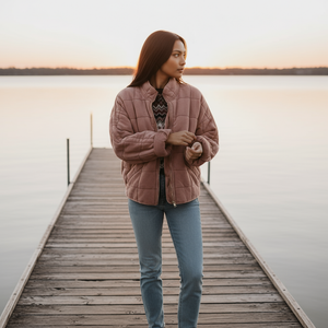 Woman standing on a wooden dock by a lake at sunset, wearing a pink quilted jacket.