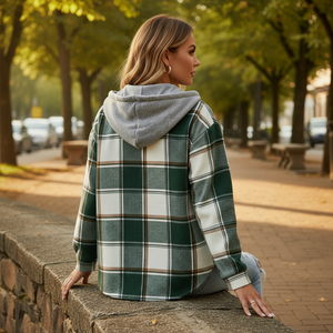 Woman wearing a plaid shirt with a hood sitting on a stone wall in an urban setting.