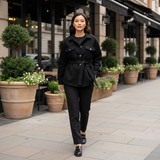 Woman in a black outfit walking on a city street with plants and buildings in the background